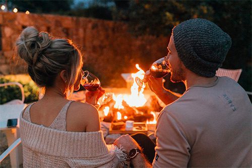 behind view of couple drinking wine, sitting by the fire-pit with lit fire.