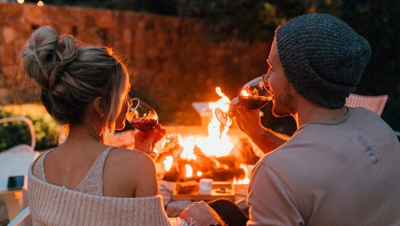 behind view of couple drinking wine, sitting by the fire-pit with lit fire.