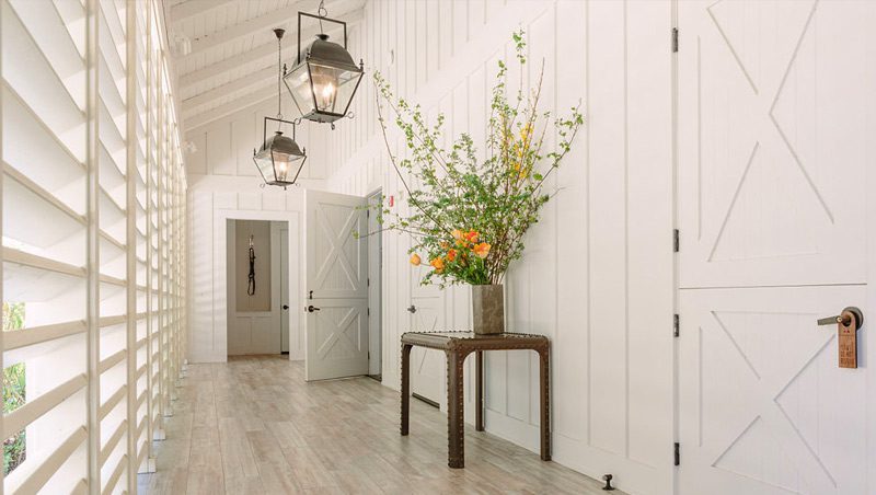 Hallway of the Farmhouse Spa. Windows with blinds on the left, wood floors, elegant lantern lights above and white wood walls.